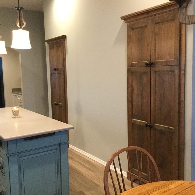 Kitchen interior with custom wooden cabinetry and pantry doors: a blue-painted island with light countertop and pendant lights, two tall stained wood built-in cabinets along a beige wall, hardwood floor and a round wooden chair.