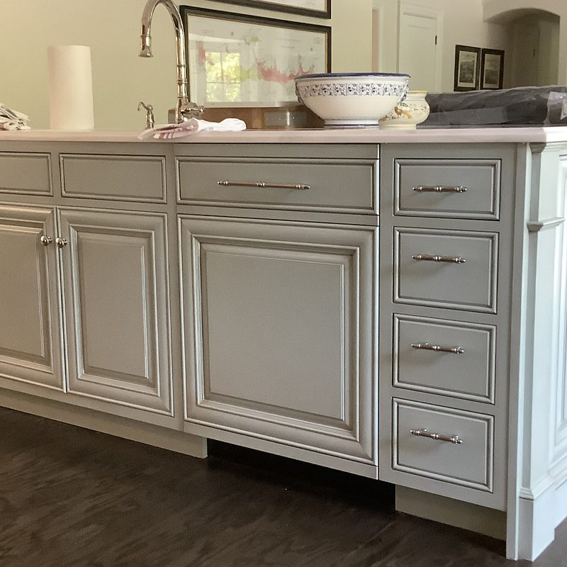 Gray painted custom kitchen cabinets and drawers with raised-panel doors, polished nickel handles and a built-in sink on dark hardwood floor, showcasing custom cabinetry and woodworking craftsmanship