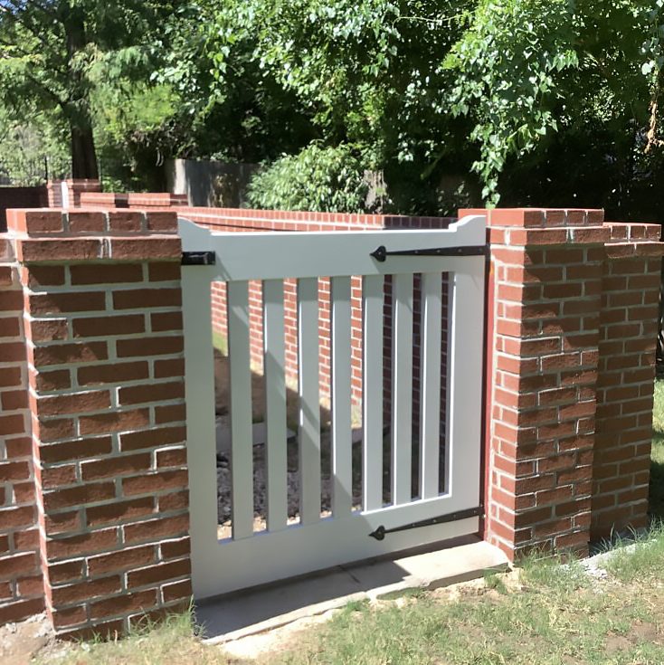 White painted wooden garden gate with vertical slats and black strap hinges set between two red brick pillars, shown outdoors with brick retaining wall and trees — custom exterior wood gate for curb appeal and cabinetry-quality woodworking.