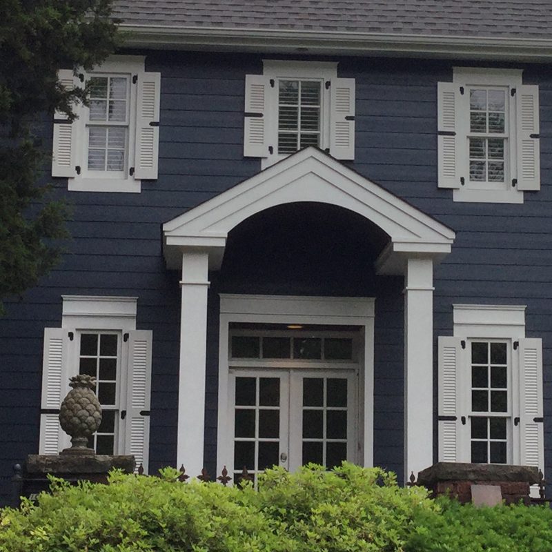 Blue two-story house front with white trim: centered entryway with pedimented portico supported by square columns, double glass doors and transom, flanked by six-over-six windows with decorative white louvered shutters, matching upper-floor windows, and low hedges in foreground
